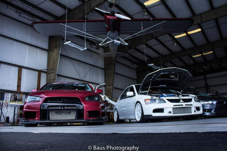 Red and white Mitsubishi Lancer Evolutions parked in an aircraft hangar, hoods up, beneath a vintage plane